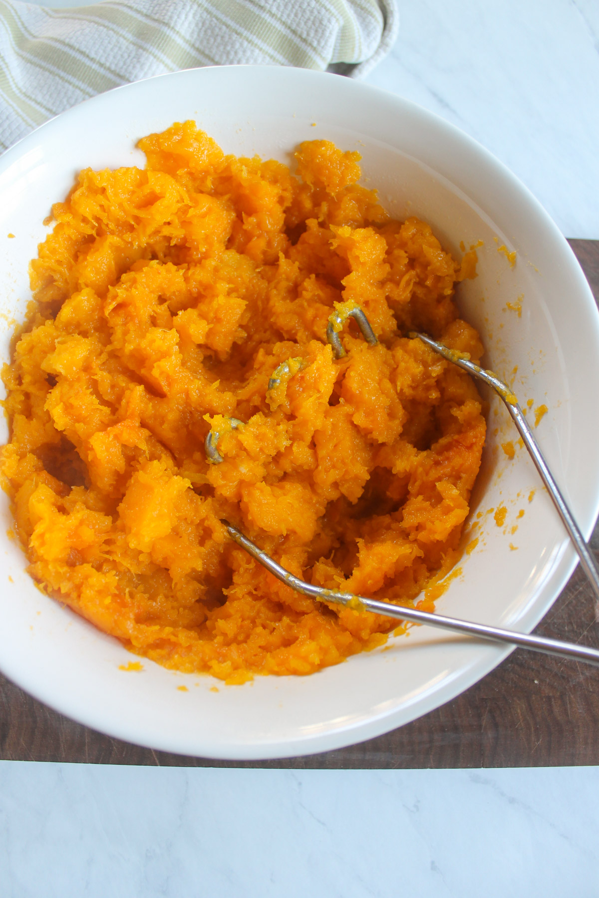 Butternut squash in a white bowl being mashed with a potato masher.