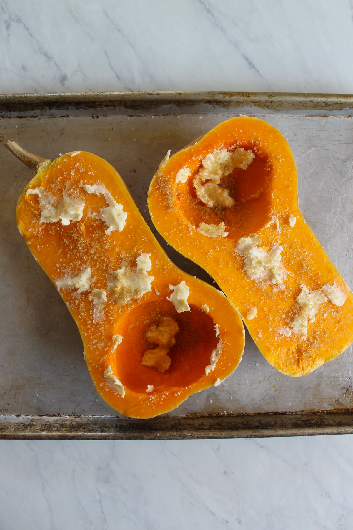 Two halves of butternut squash on a sheet pan smeared with butter, brown sugar and salt.