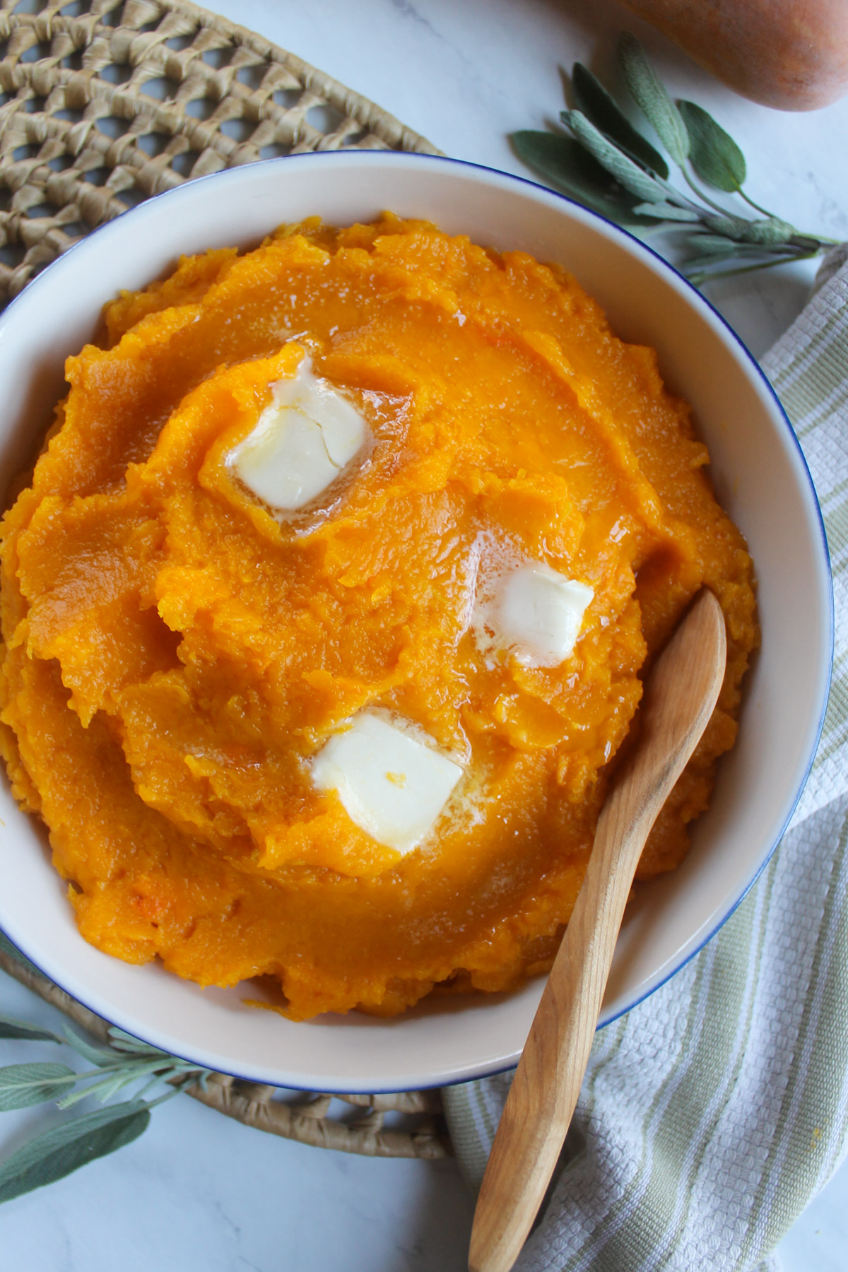 A bowl of mashed butternut squash topped with butter on a wicker mat with sage leaves.