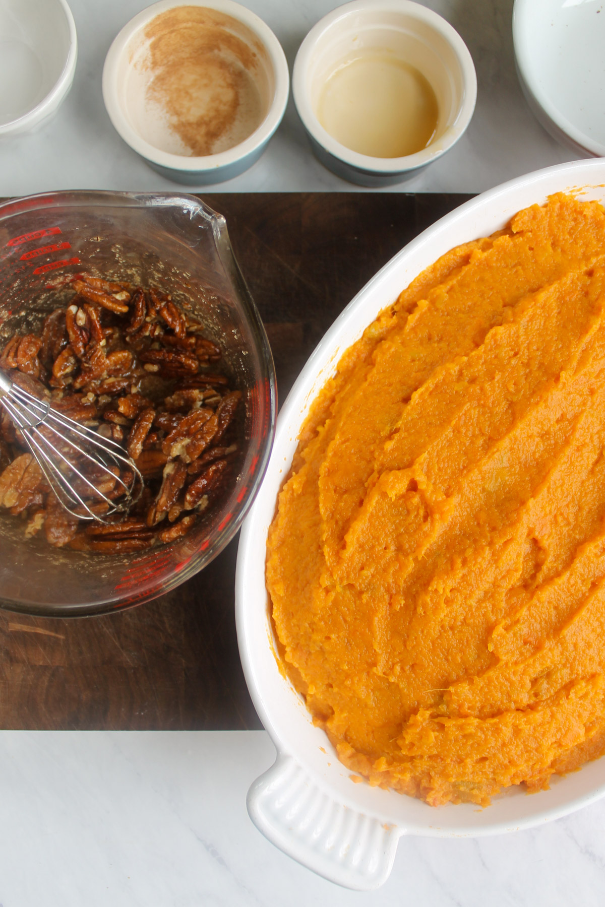 Mashed sweet potatoes in a baking dish and a glass measuring cup of maple pecan topping with a whisk.