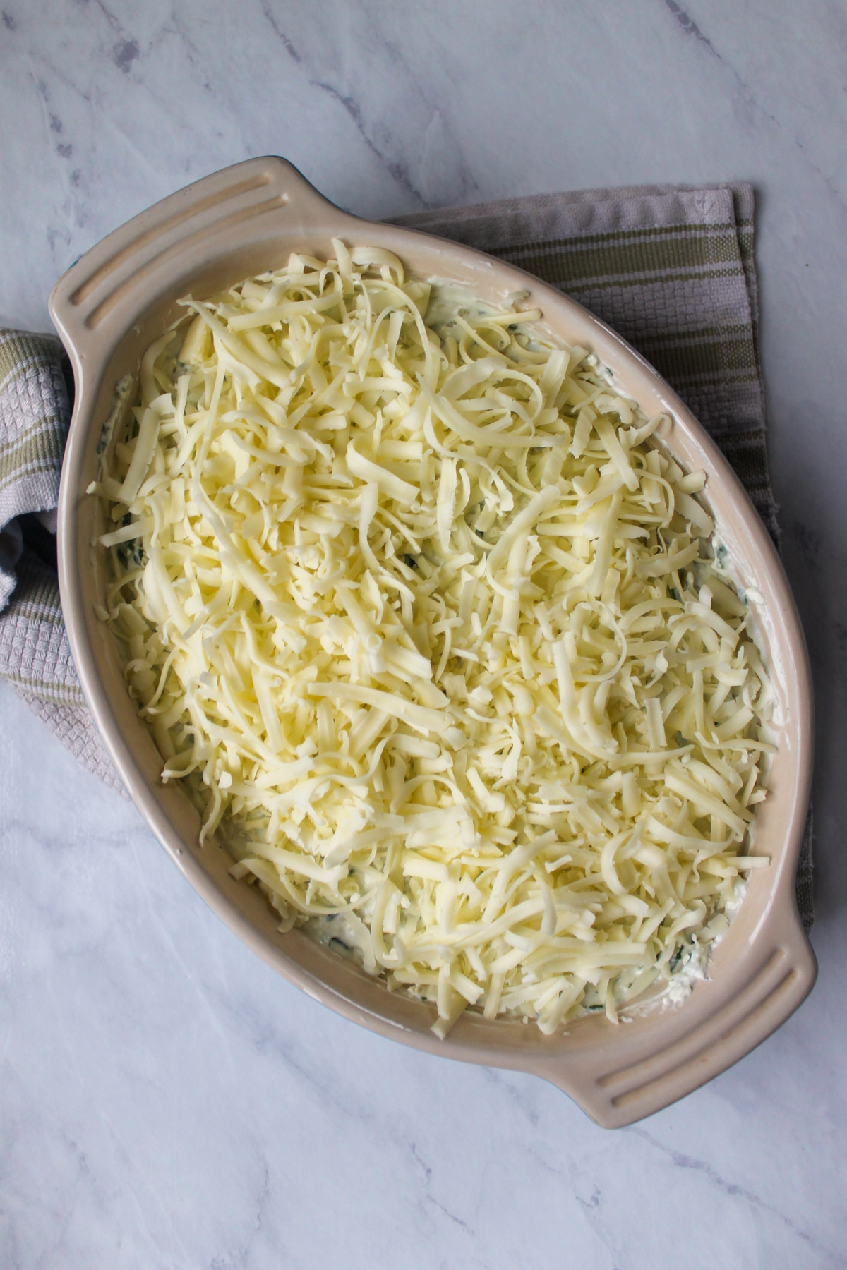An oval baking dish with the artichoke dip and shredded white cheddar cheese ready to bake.
