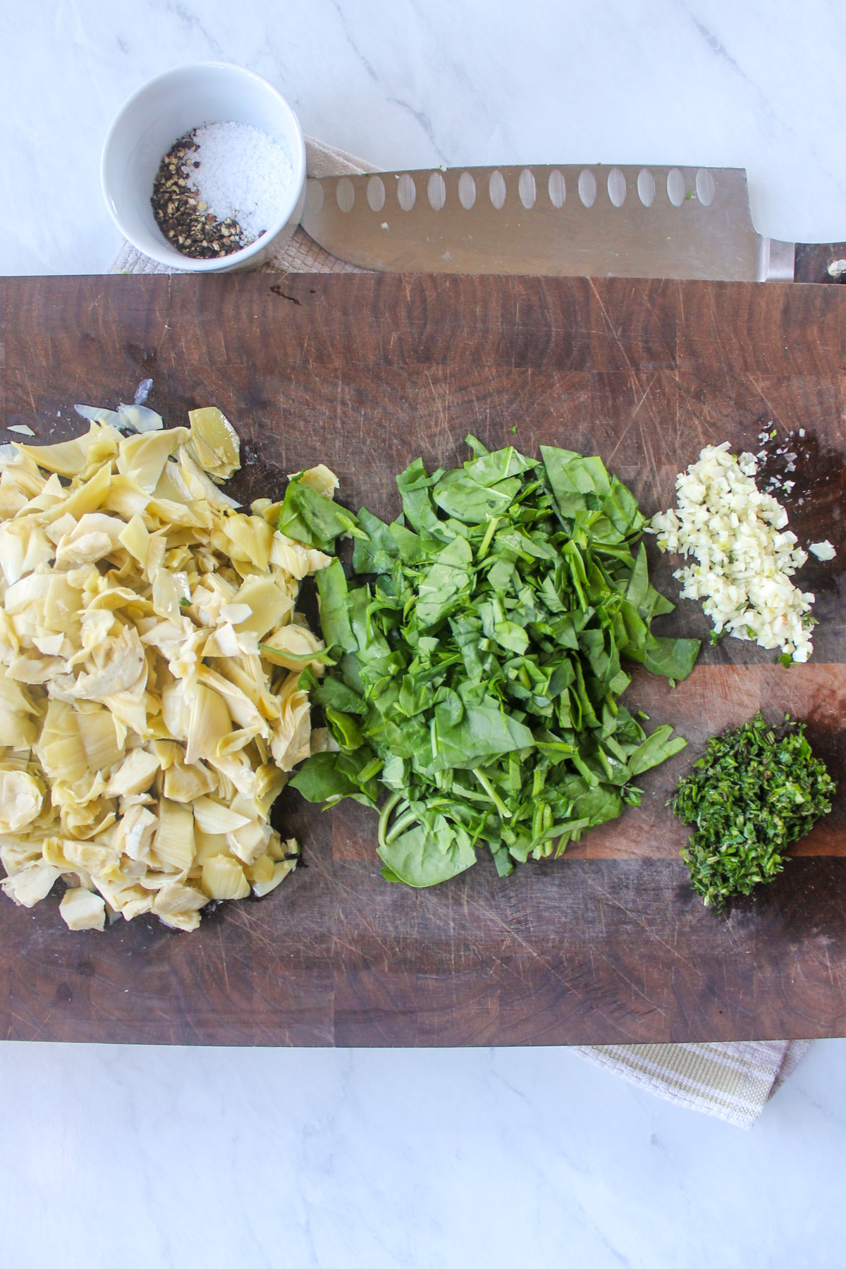 Chopped artichokes, fresh spinach, herbs and garlic on a wooden cutting board.