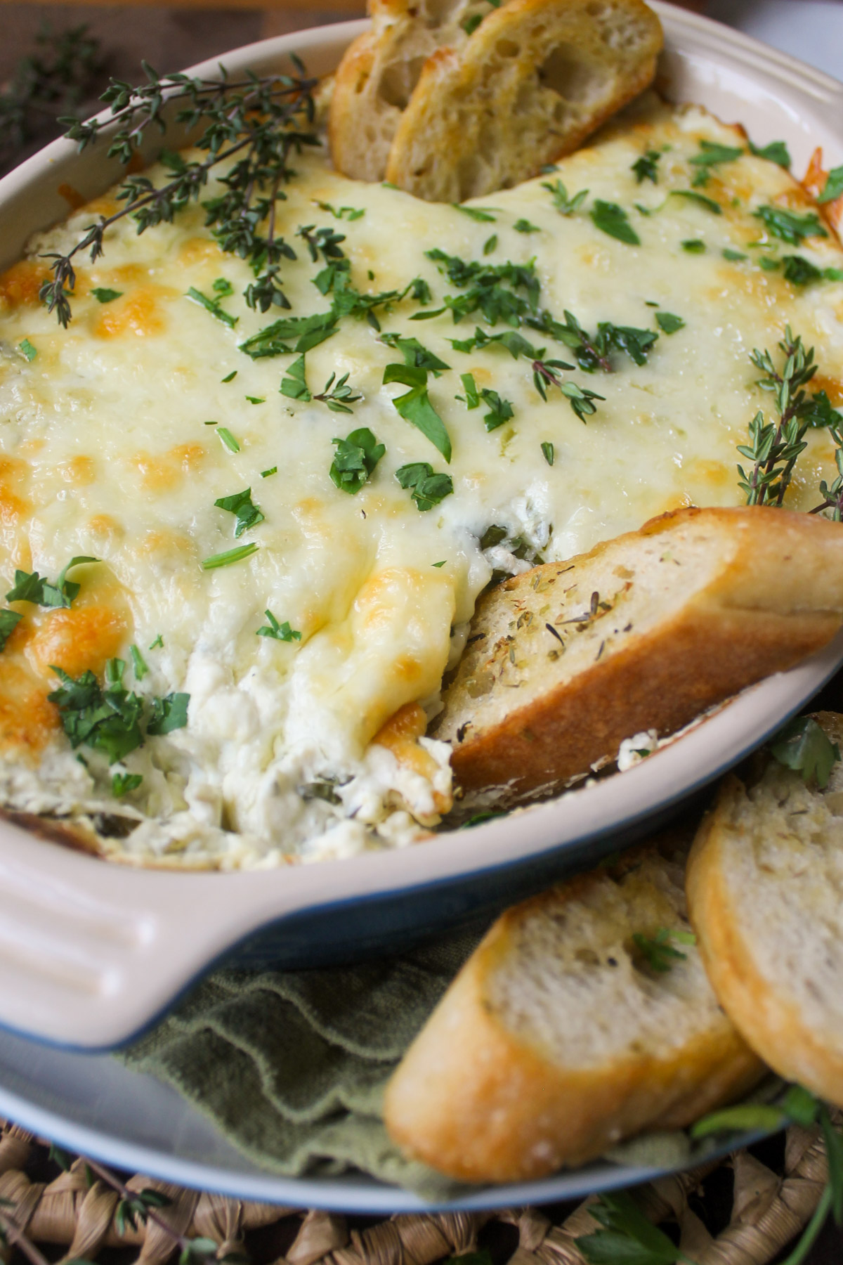 An oval baking dish of goat cheese artichoke dip with herbs and bread for dipping.