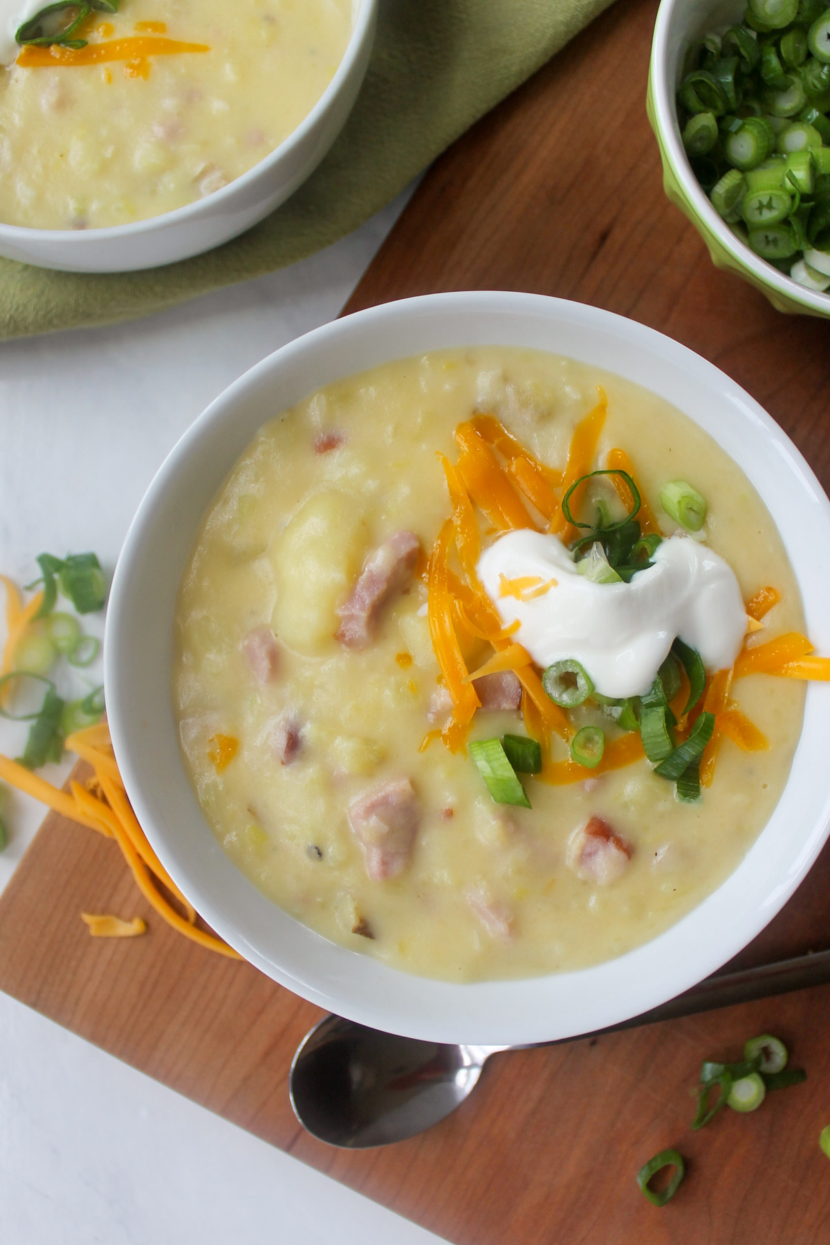 A white bowl of Potato Ham and Leek Soup with cheese, plain yogurt and sliced scallions.