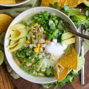 A white bowl of white chicken chili with green chopped herbs, avocado slices, and tortilla chips.