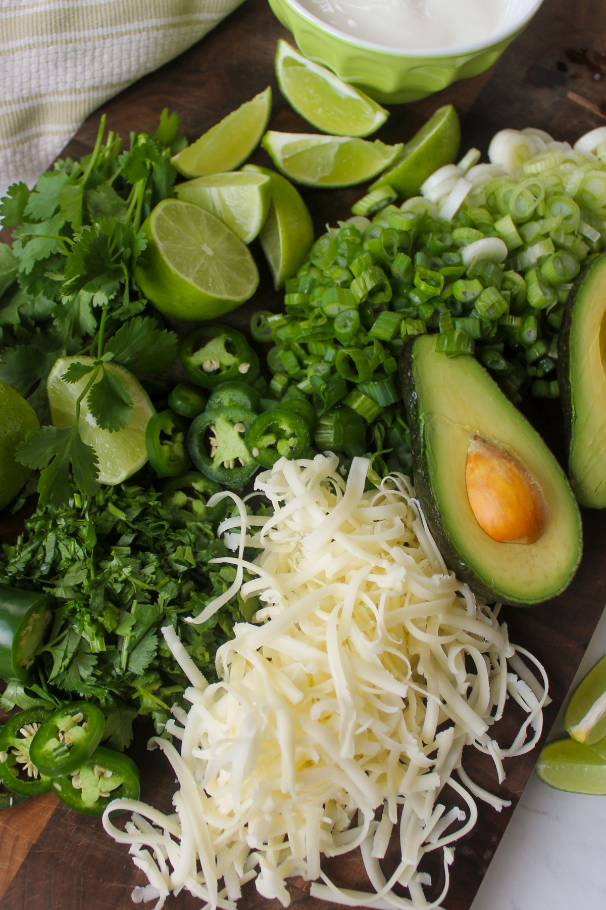 A cutting board with chili toppings like avocado, white shredded cheese, lime wedges, scallions, cilantro and jalapeno.