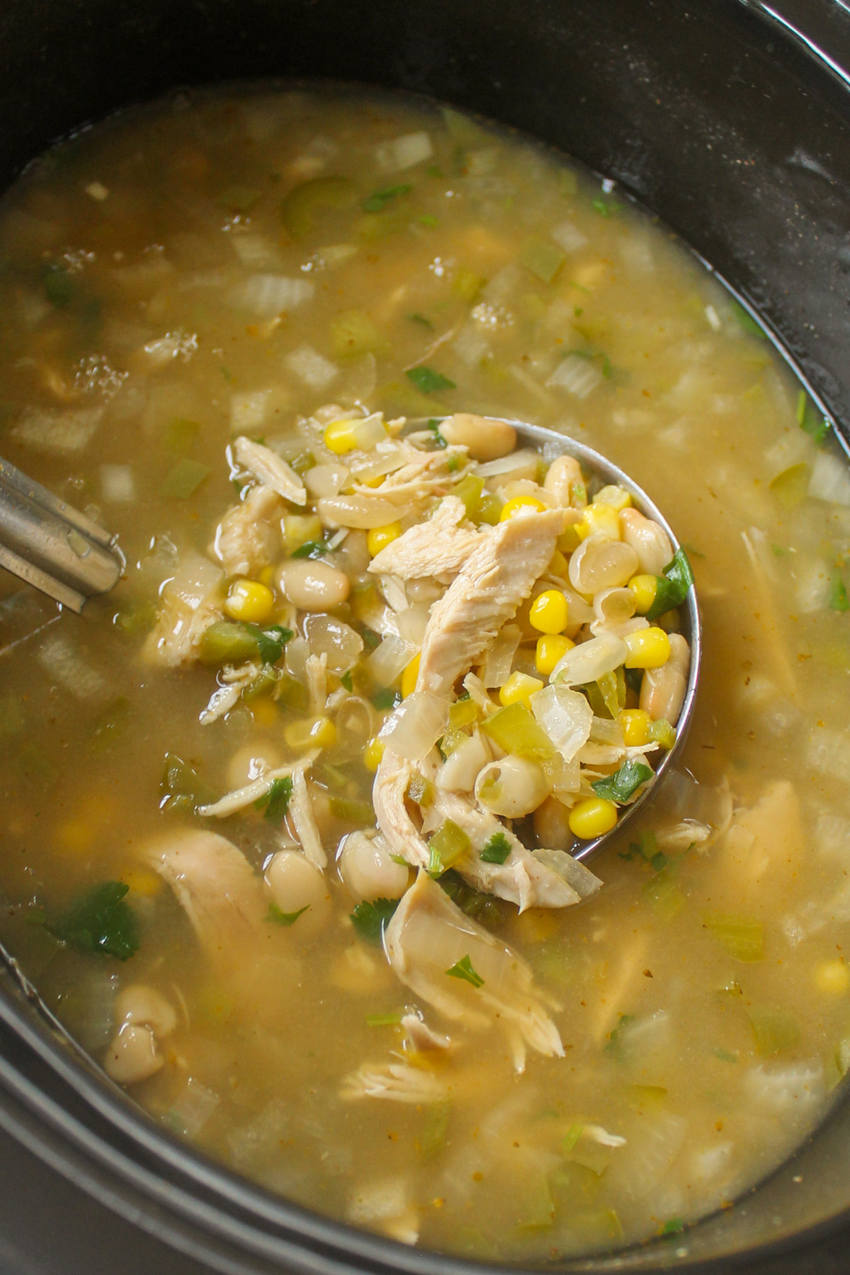 A slow cooker of white chicken chili with a ladle showing the shredded chicken, corn and broth.