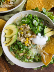A bowl of white chicken chili with sliced jalapeno, cheese, yogurt, avocado and tortilla chips.