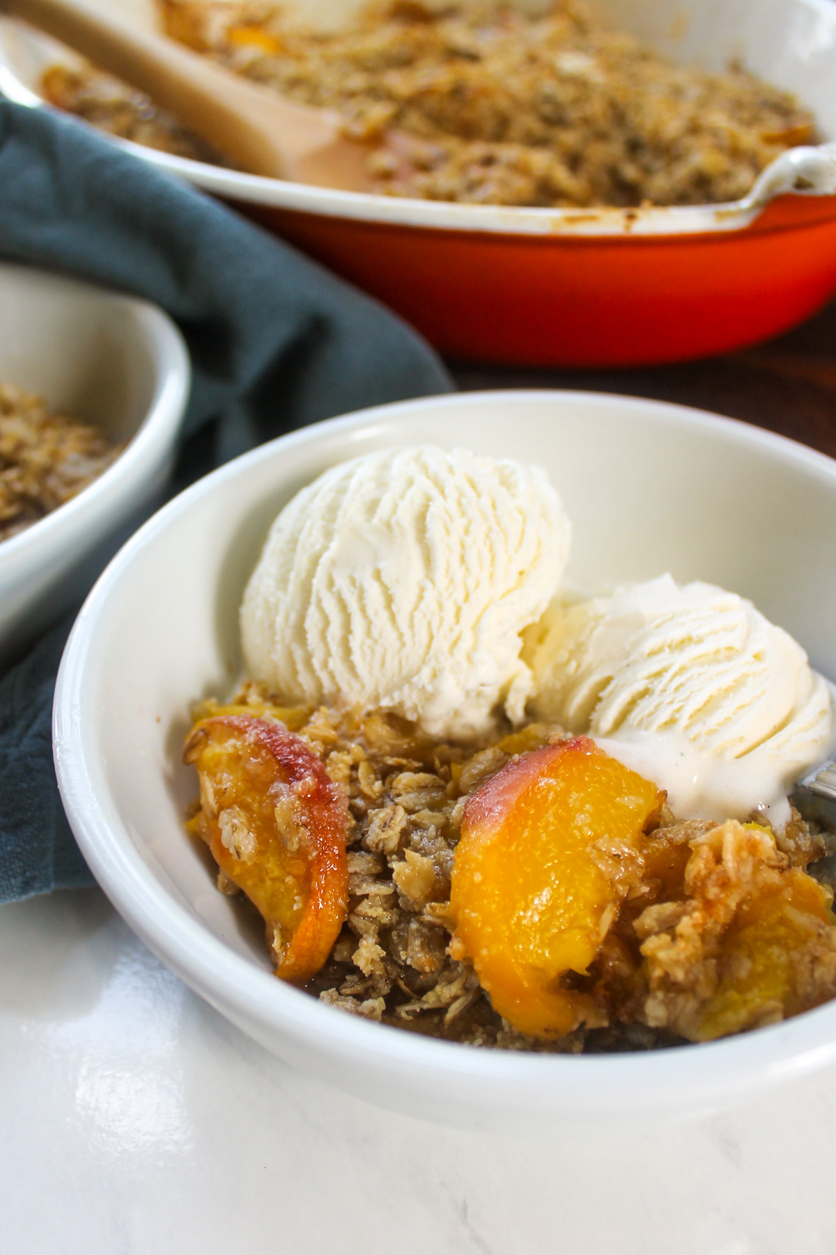 A serving of peach crisp with ice cream in front of the baking dish.