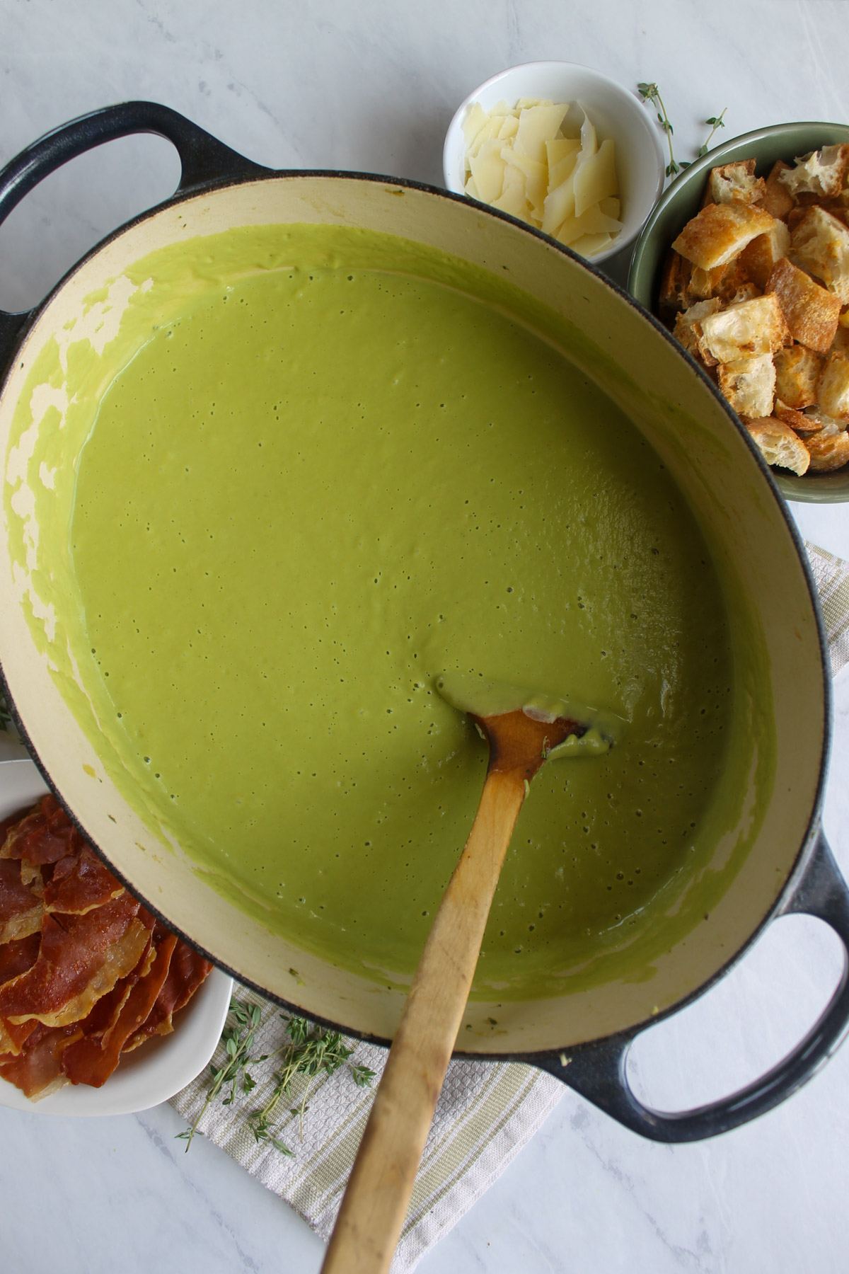 A large soup pot of pureed Asparagus Broccoli Soup with bowls of croutons, crispy prosciutto and Parmesan cheese.