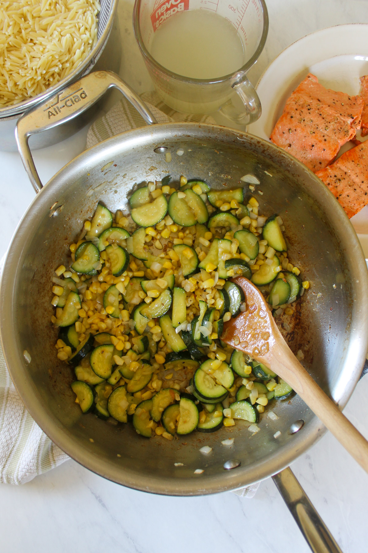 Sauteed zucchini and corn in a skillet next to a plate of cooked salmon filets.