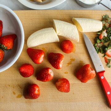 Slicing a banana and cutting the tops off strawberries on a cutting board.