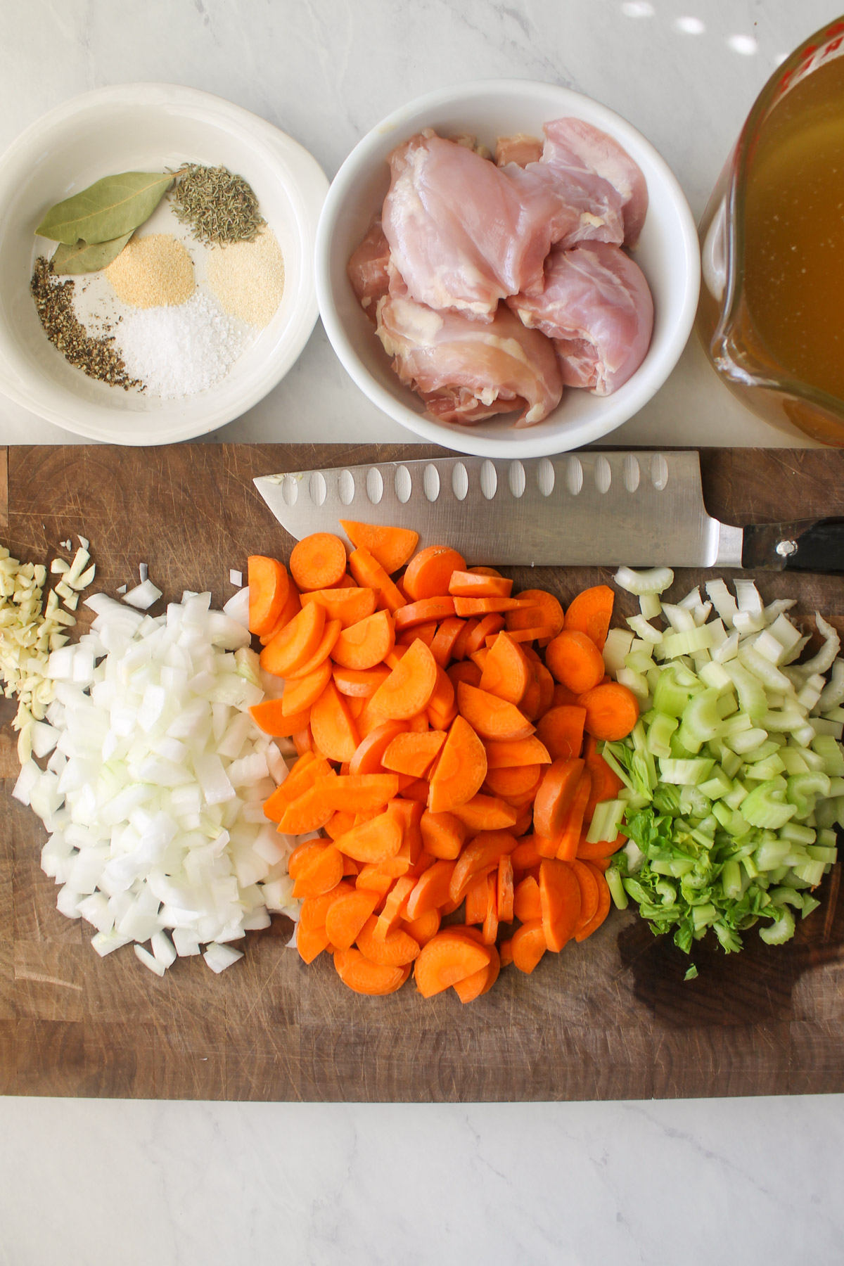 Chopped onion, carrots, celery and garlic on a cutting board with a bowl of raw chicken and seasonings.