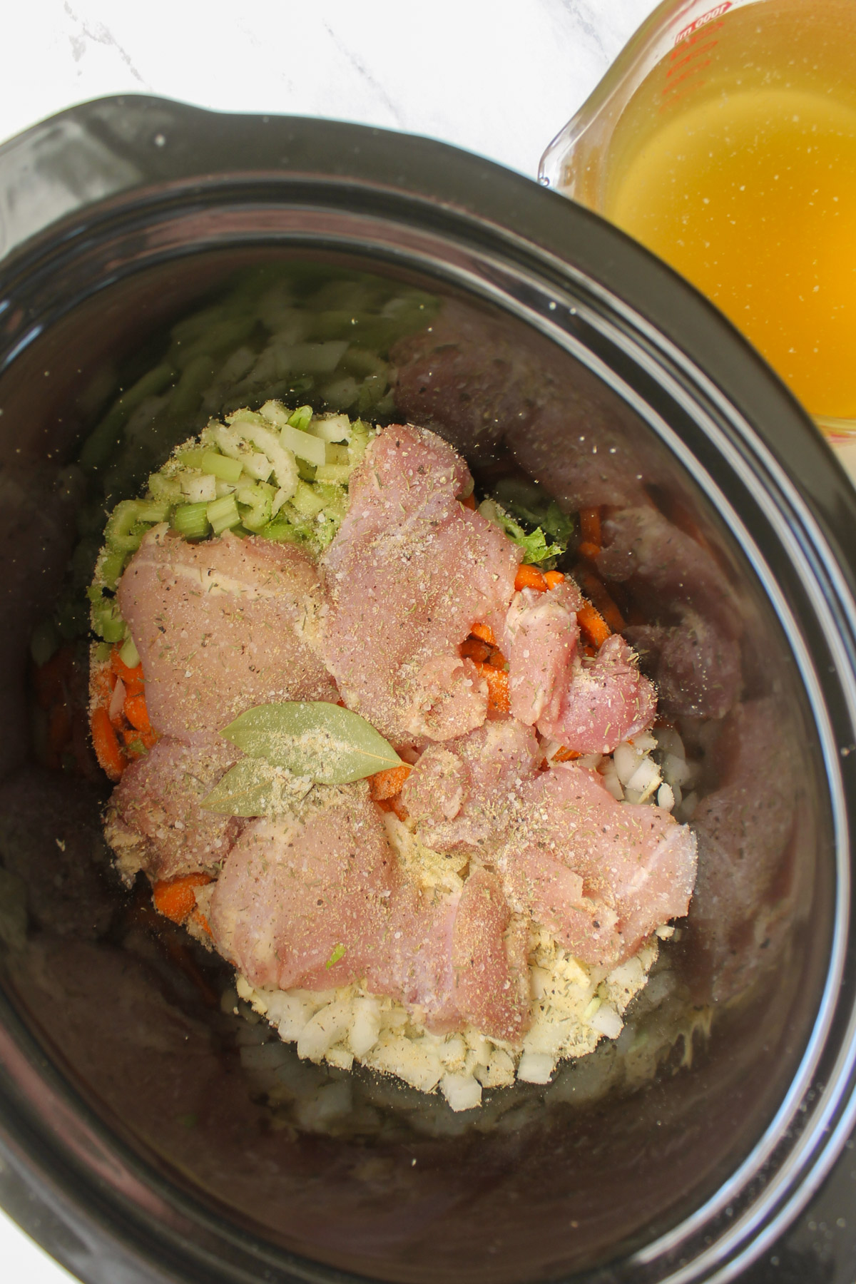 A crockpot with vegetables, raw chicken and seasoning.