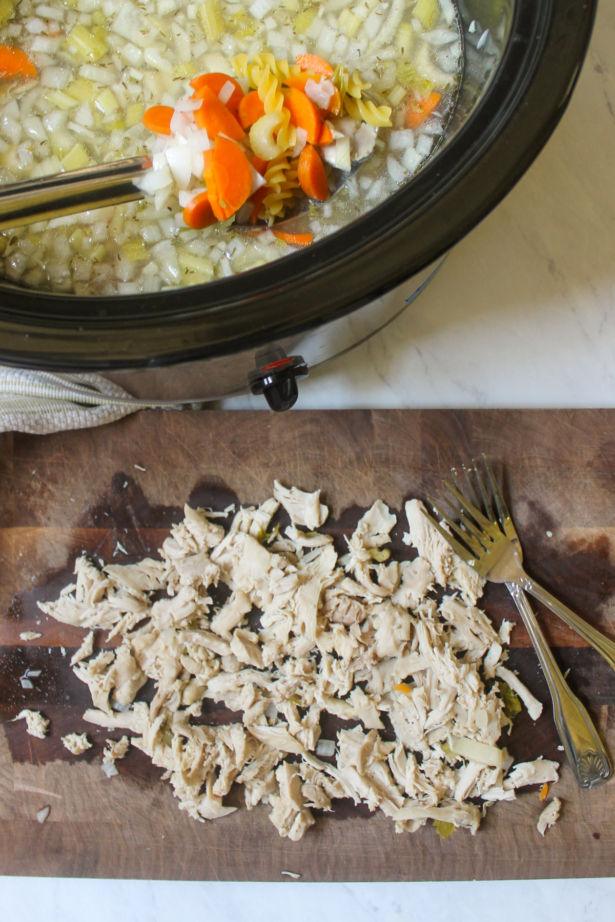 Chicken shredded with forks on a cutting board next to a crockpot of chicken noodle soup.