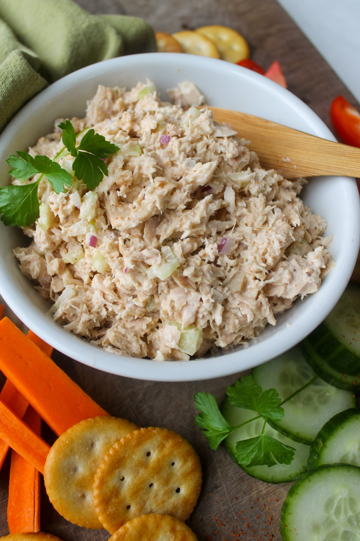 A bowl of tuna salad on a cutting board with cucumber, carrots and crackers.