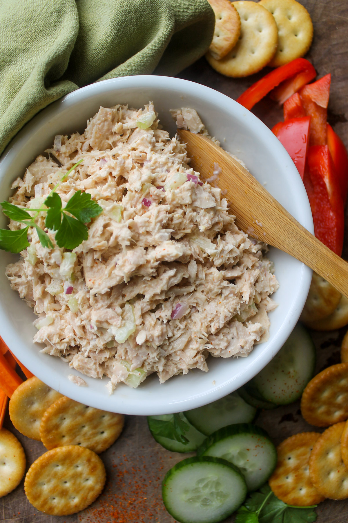 A white bowl of tuna salad surrounded with red bell peppers, cucumbers, and crackers.