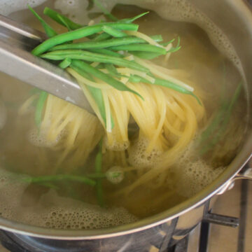 Pasta and green beans boiling together in a pot lifted with tongs.