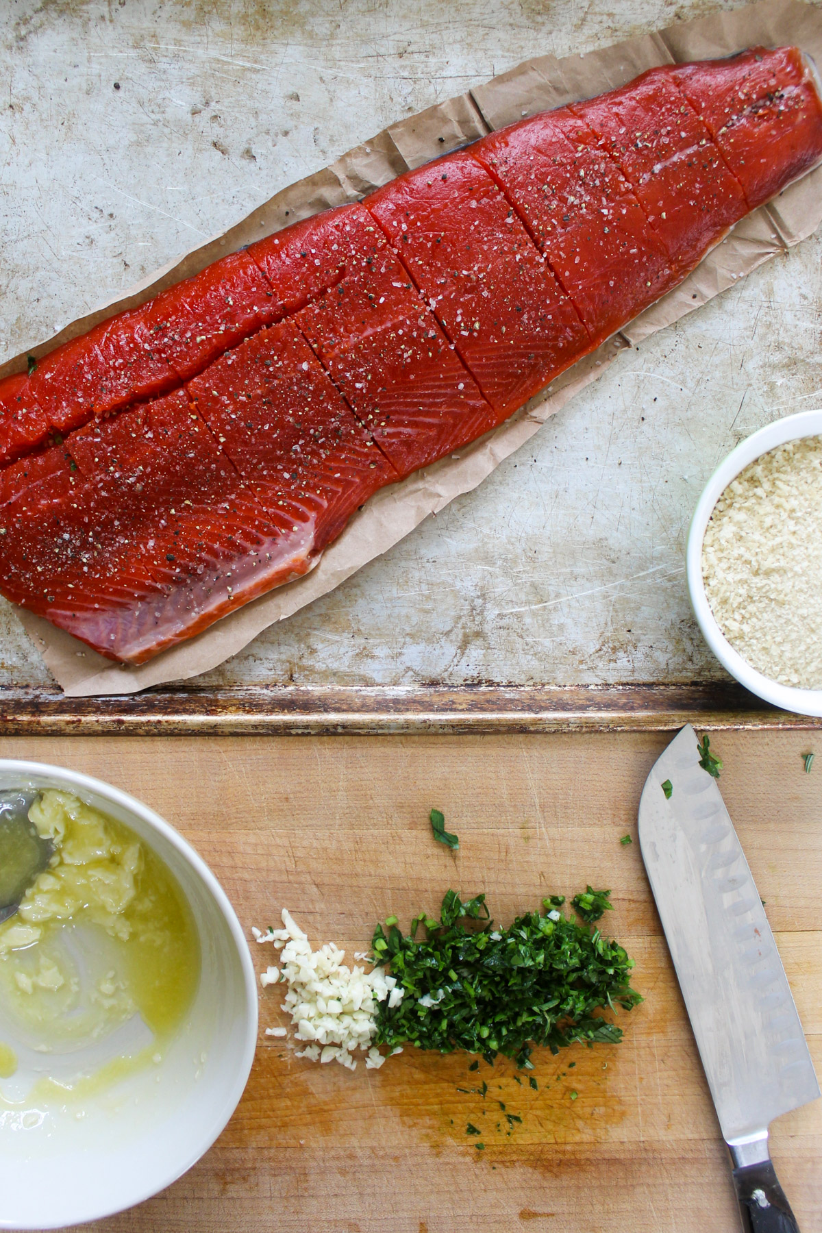 Fresh garlic and herbs chopped on a cutting board with a raw filet of salmon on a sheet pan.