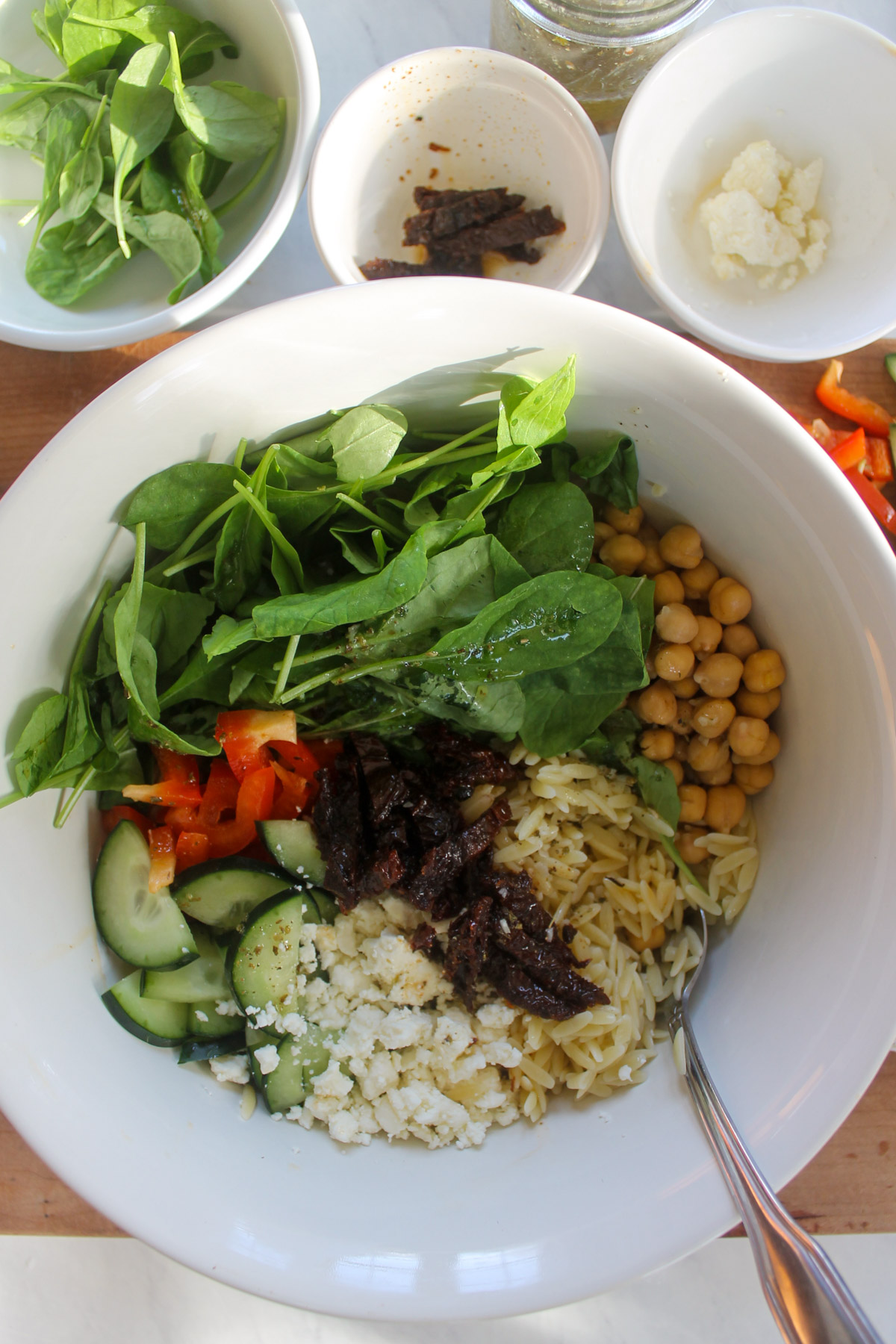 A bowl of salad ingredients like orzo, chickpeas, arugula, peppers, feta, cucumber and sun-dried tomato.