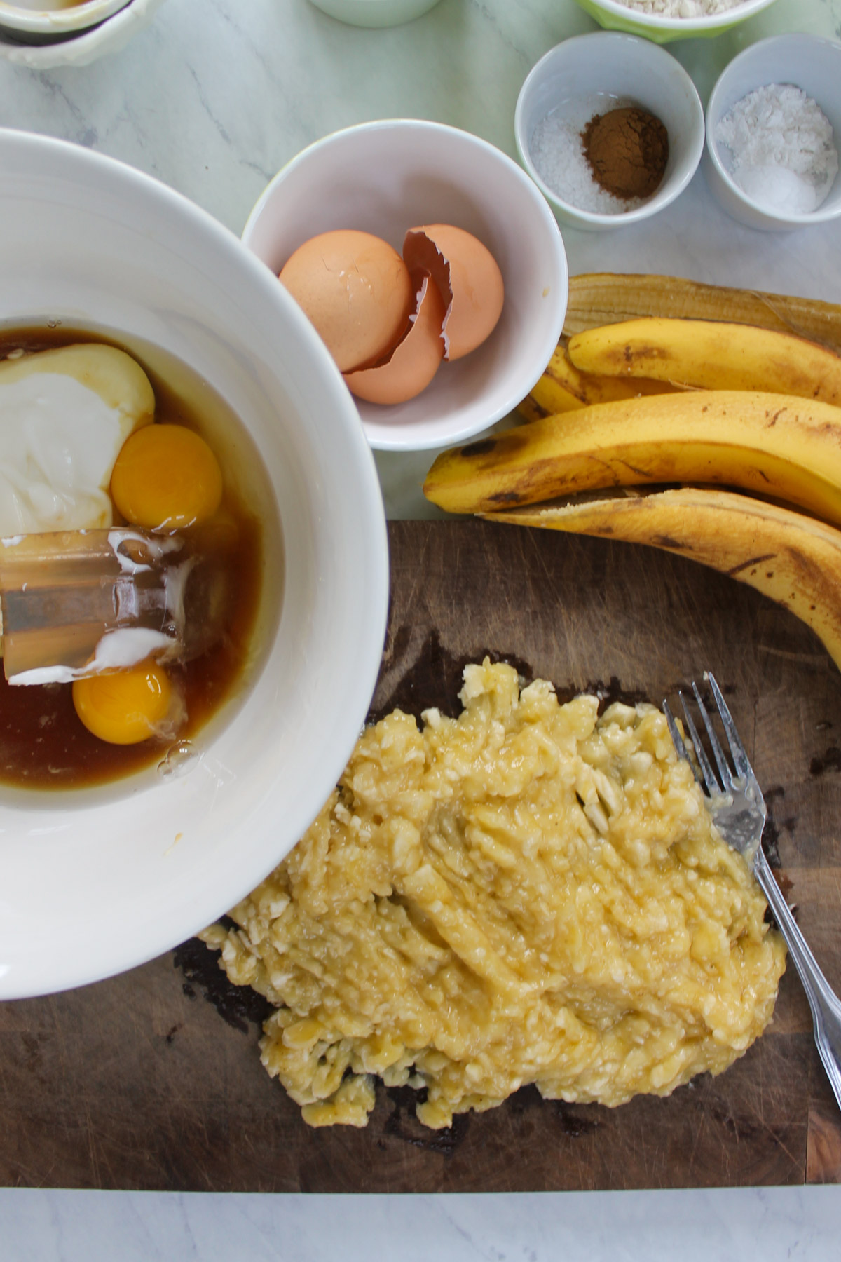 Mashed banana on a cutting board with a bowl of eggs, yogurt and maple syrup.