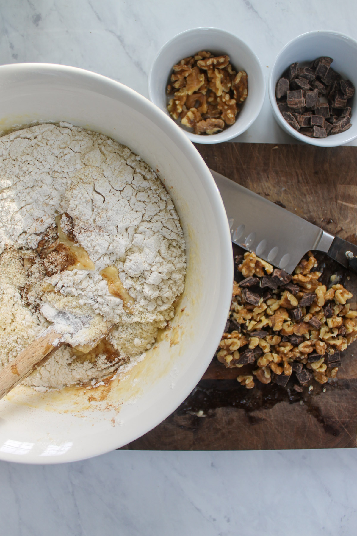 Adding dry ingredients to a bowl of banana bread batter and chopping walnuts and chocolate.