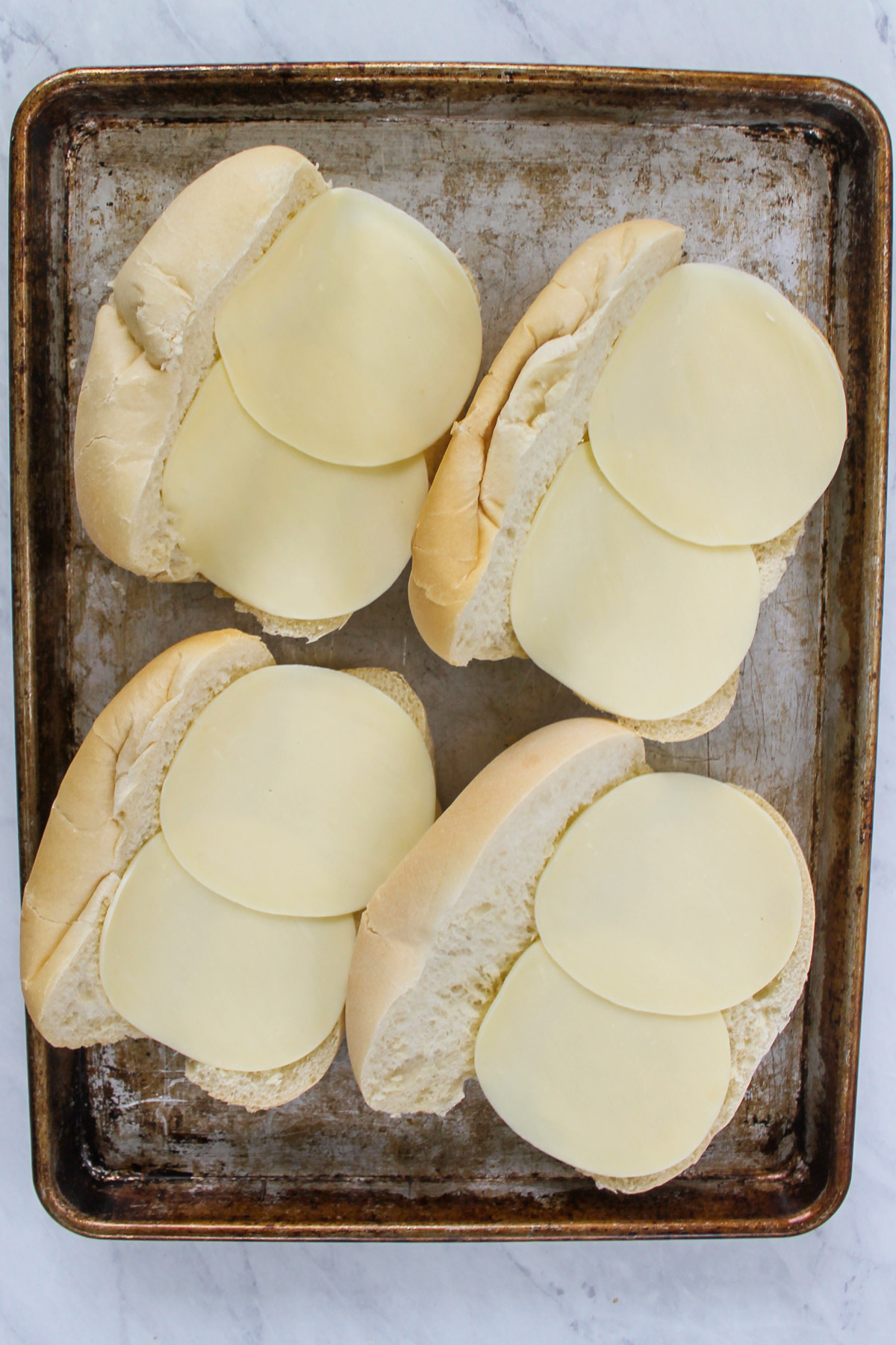 Four hoagie buns on a sheet pan with sliced provolone cheese.