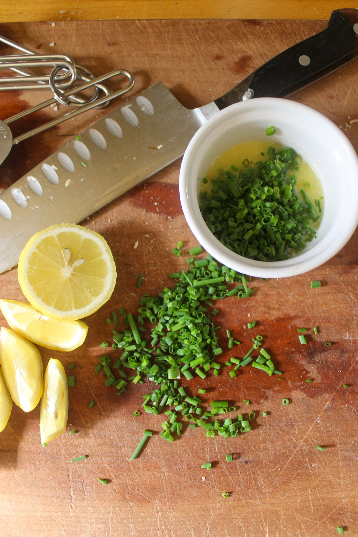Chopped chives on a cutting board with melted butter and lemon wedges.
