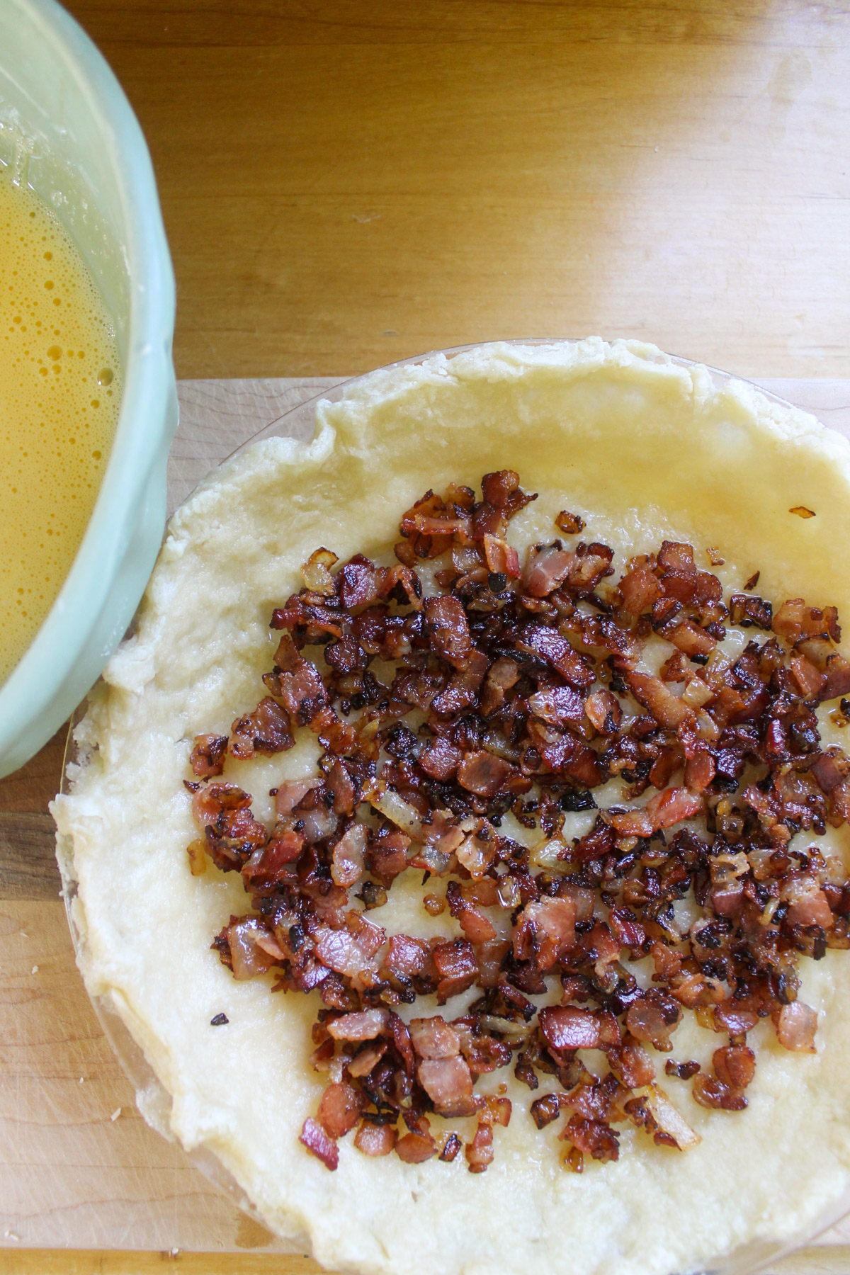 Cooked bacon and onion in a pie crust with a bowl of eggs.