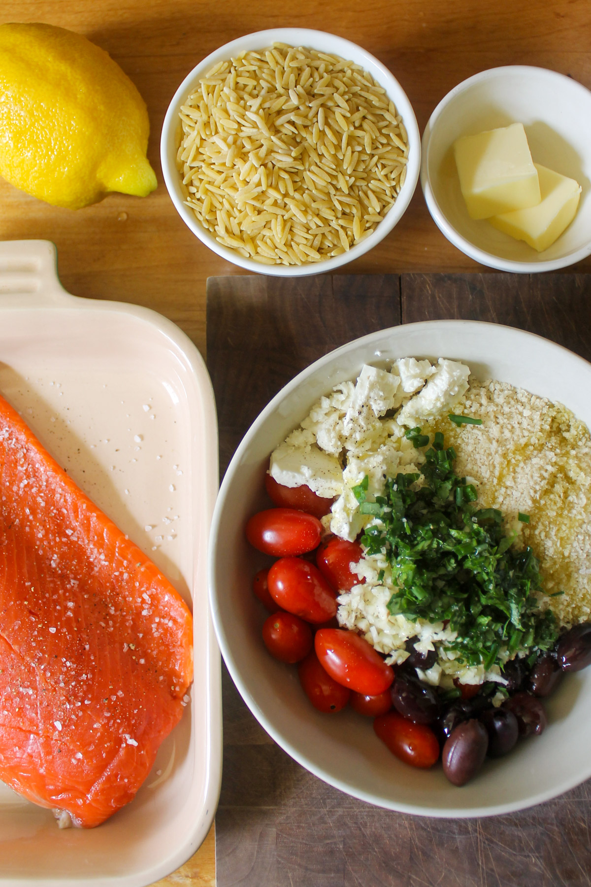 A filet of raw salmon in a baking dish and a bowl of topping with feta, tomato, herbs, panko and olives.