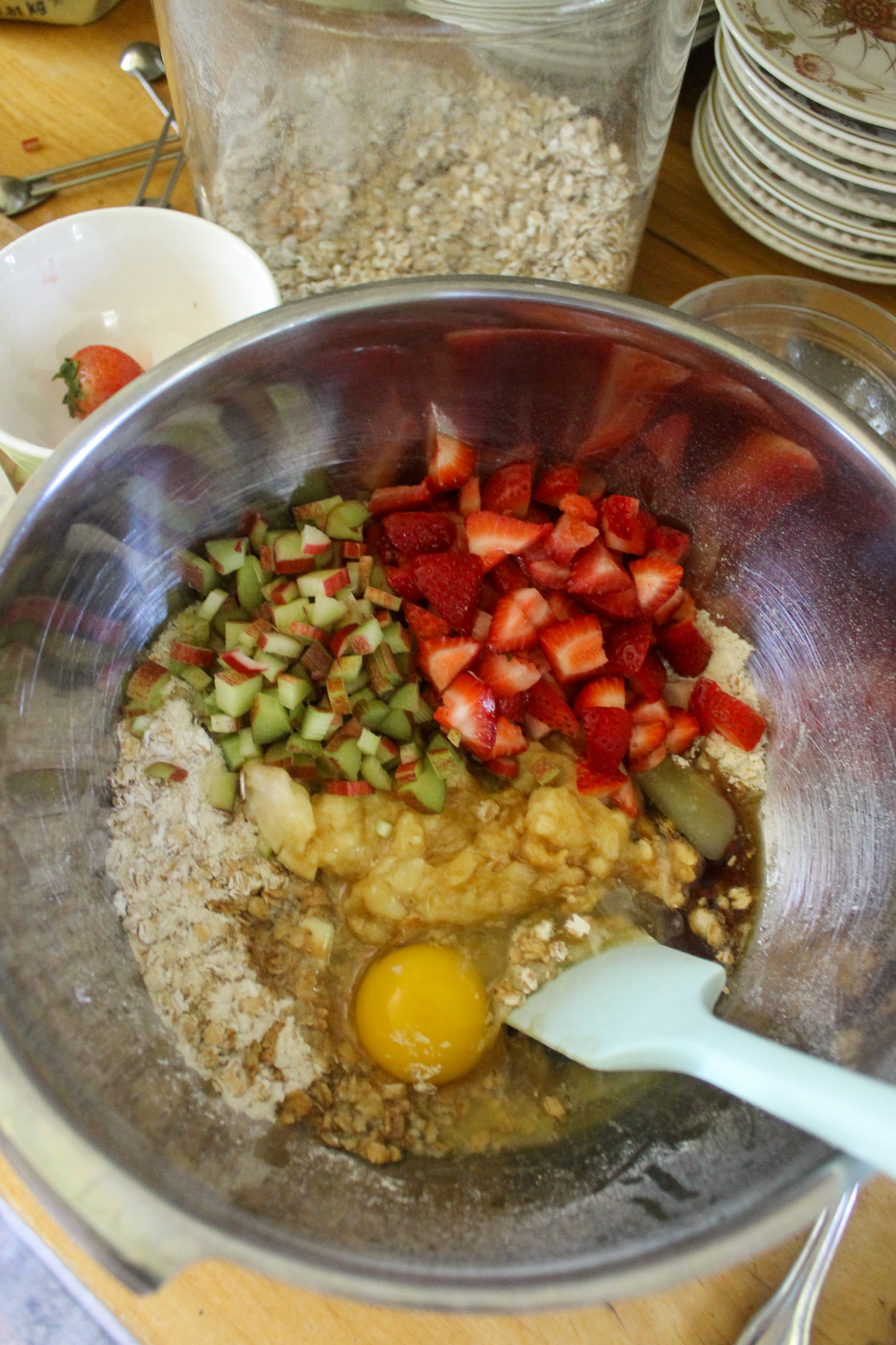 Adding strawberries, rhubarb, eggs, and mashed banana to the bowl of muffins batter.