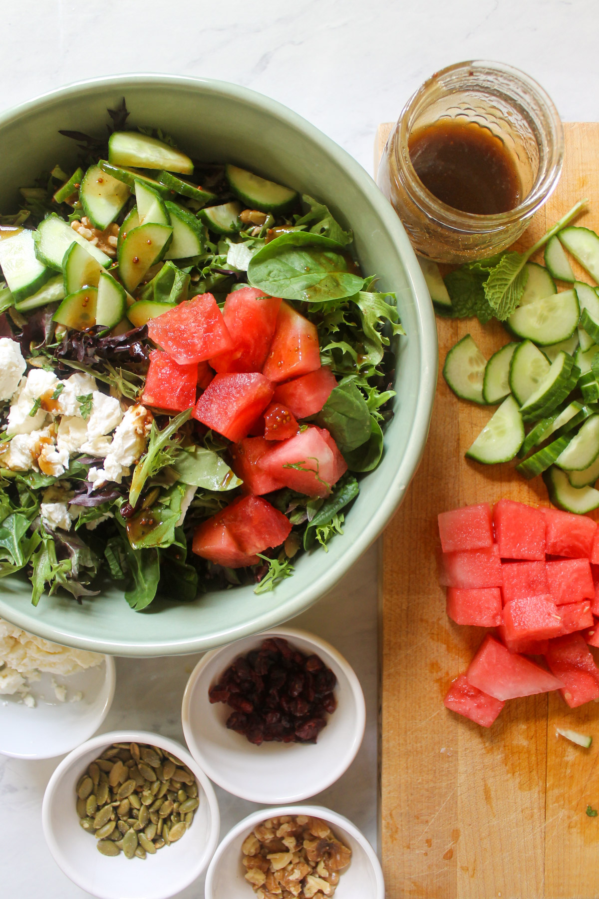 Adding lettuce, cucumber, watermelon, feta cheese, walnuts, dried cranberry, pumpkin seeds and dressing to a salad bowl.