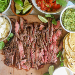 Sliced flank steak for tacos with bowls of toppings and tortillas.