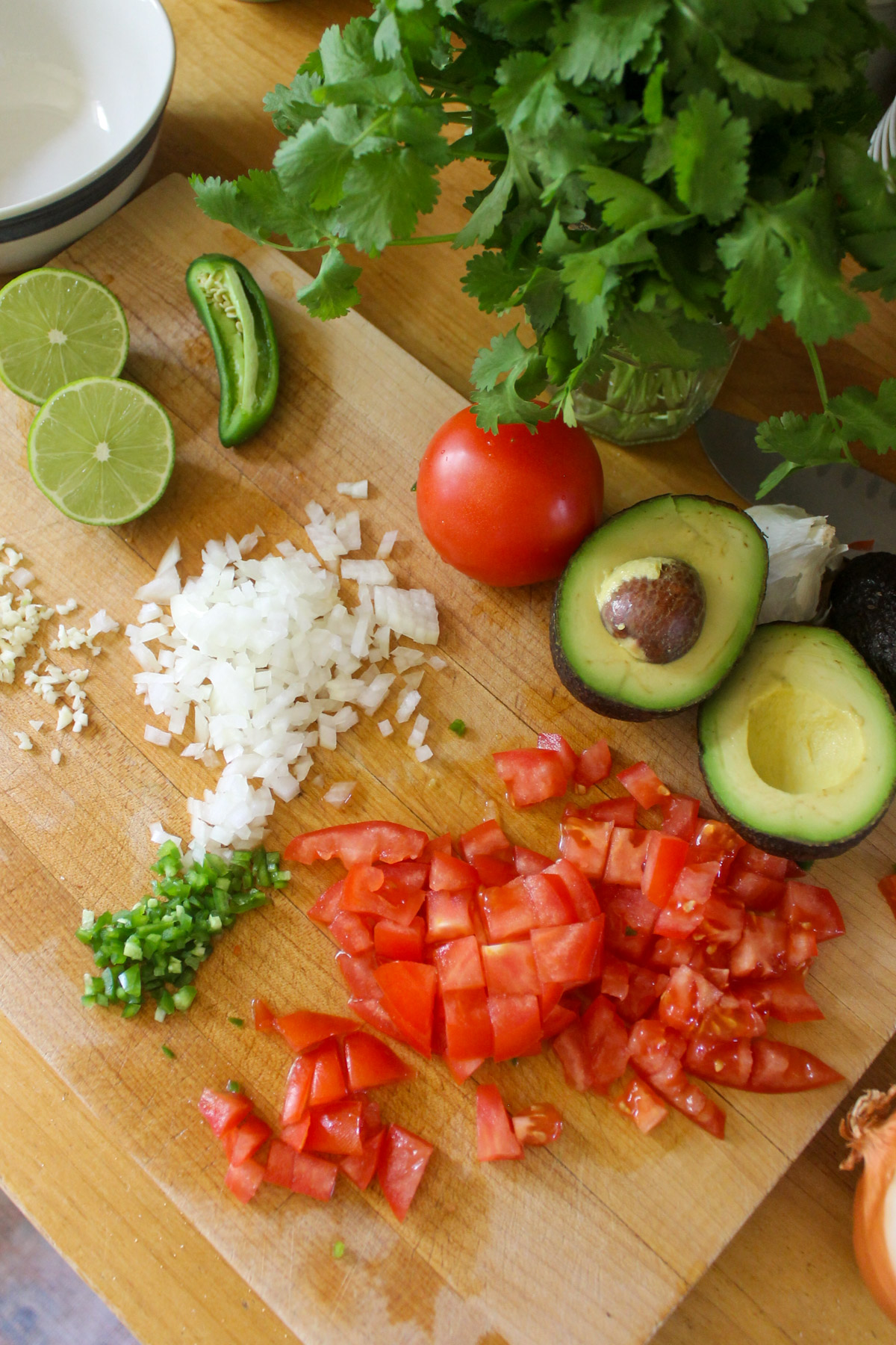 Chopped tomato, onion, garlic, jalapeno and avocado on a cutting board with cilantro and lime.