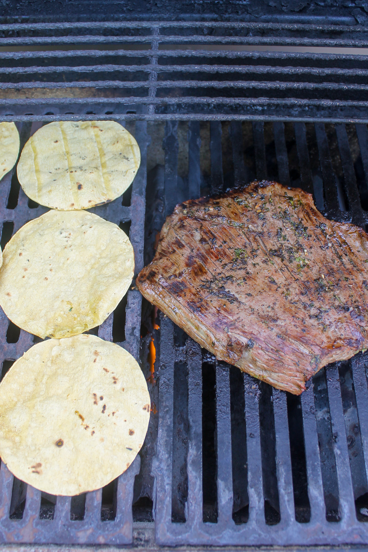 Grilled flank steak next to corn tortillas on a grill.