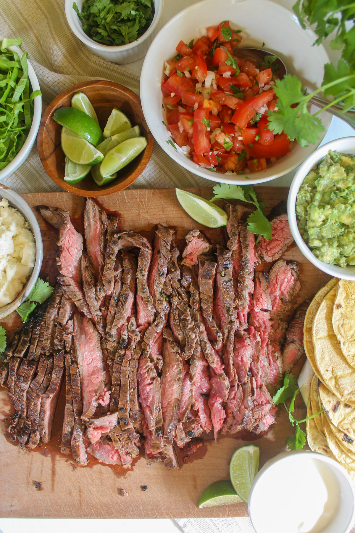 Sliced flank steak on a cutting board with taco toppings like pico de gallo, guacamole, limes and cilantro.