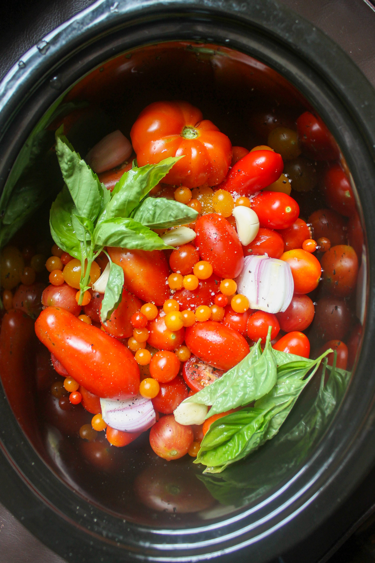 Raw whole tomatoes in a slow cooker with onions, garlic cloves and fresh basil.